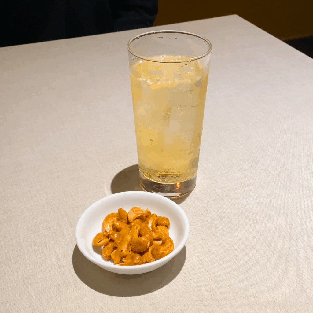 一人分のドリンクとつきだしが置かれたテーブル。ほろ酔い壺中夜を注文したら付いてくるスパイシーカシューナッツはやみつきになるおいしさを表現