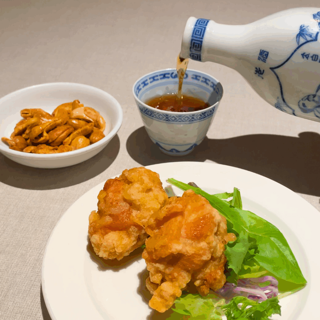 スパイシーカシューナッツと鶏の唐揚げを並べたテーブルに、紹興酒を注ぐシーン。ほろ酔い壺中夜のつきだしとおつまみのイメージ。