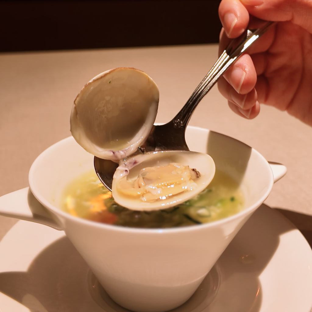 貝付きのハマグリをスプーンですくい上げたシーン。青海苔の香りが漂う優しい色合いのスープで、春の訪れを思わせる上品な中華スープ料理。