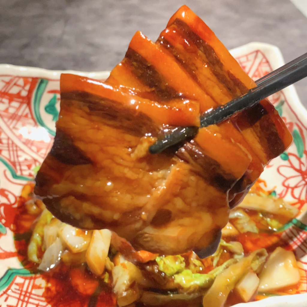 お箸で持ち上げられた豚バラ肉のアップ。柔らかく煮込まれた肉とタレの光沢が際立つ