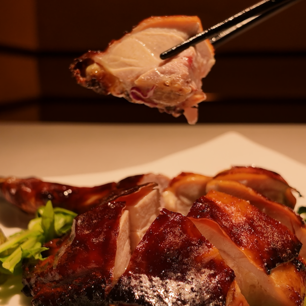 クリスピーチキンの一切れを箸で持ち上げ、カリカリの皮の照りとジューシーな断面が見えるアップ写真。スパイスの香りと食感の良さが伝わる料理カット。